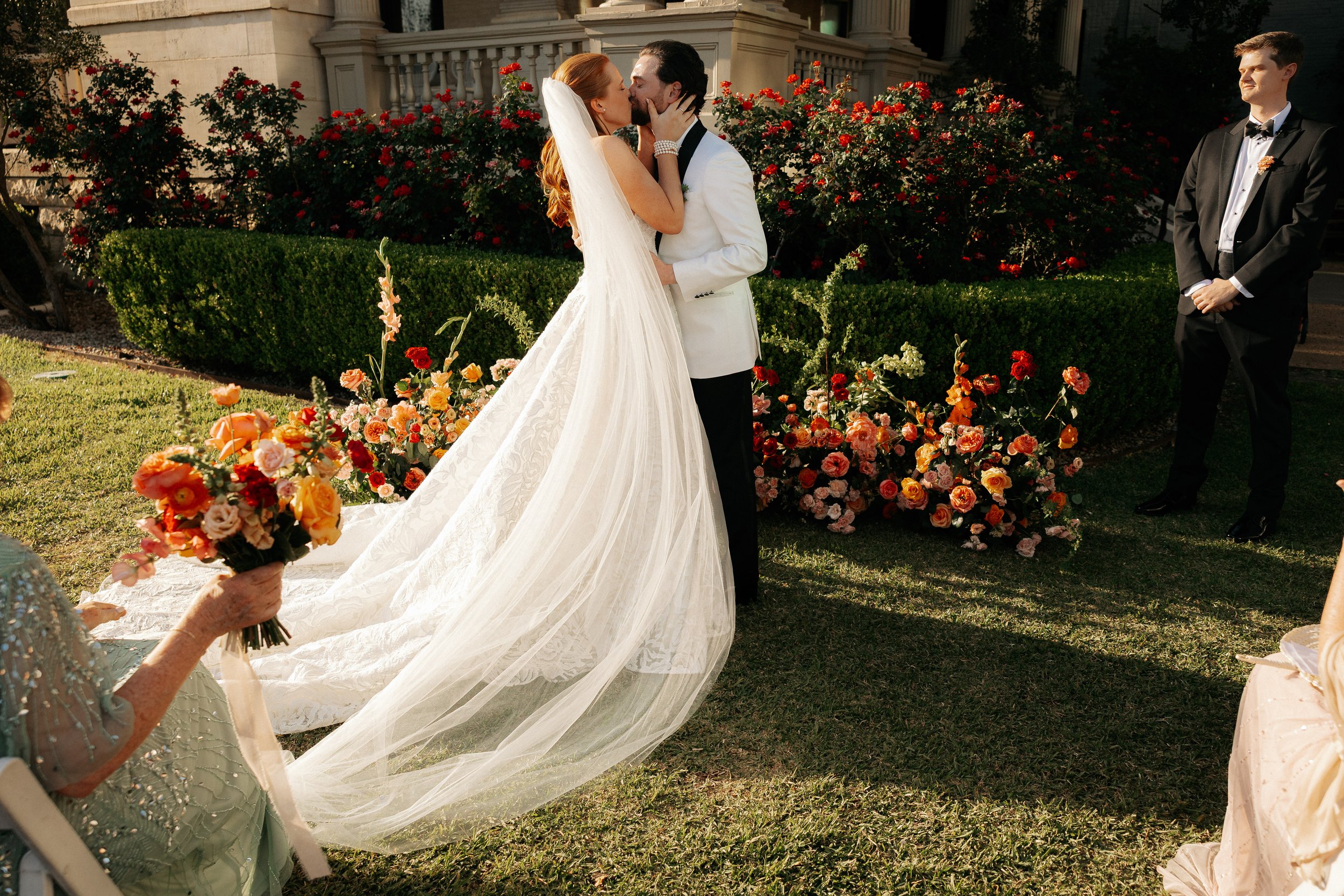 A bride in a long veil kisses a groom in a white dinner jacket on a sunlit garden lawn